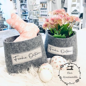 Könnte beinhalten: Zwei graue Filz-Körbe mit dem Text "Frohe Ostern" darauf. Die Körbe sind mit rosa Blumen und einem rosa Hasen gefüllt. Es gibt auch drei weiße Ostereier mit Blumenmustern.