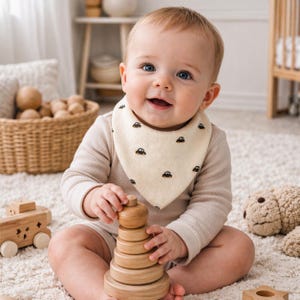Pode incluir: Um bebê sorrindo usando um babador creme com ilustrações de carros pretos. O bebê está brincando com um brinquedo de empilhamento de madeira. Outros brinquedos de madeira e uma cesta de vime estão no fundo. O bebê está usando um body bege de manga comprida.