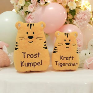 Op de afbeelding: Twee pluche tijger speelgoed met geborduurde gezichten en strepen. De grotere tijger is geel met de tekst "Troost Kumpel" geborduurd op de voorkant. De kleinere tijger is geel met de tekst "Kraft Tigerchen" geborduurd op de voorkant.