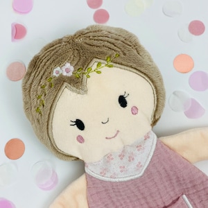 K&ouml;nnte beinhalten: Eine weiche Pl&uuml;schpuppe mit braunen Haaren, einem rosa Kleid und gestickten Gesichtsz&uuml;gen. Die Puppe tr&auml;gt ein Blumenband und ist von Konfetti umgeben.