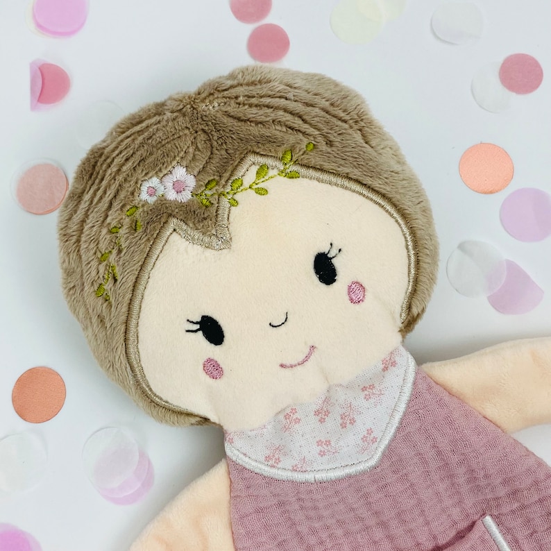 K&ouml;nnte beinhalten: Eine weiche Pl&uuml;schpuppe mit braunen Haaren, einem rosa Kleid und gestickten Gesichtsz&uuml;gen. Die Puppe tr&auml;gt ein Blumenband und ist von Konfetti umgeben.