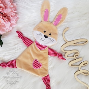 Può includere: Una coperta di sicurezza a forma di coniglio beige con orecchie rosa e braccia e gambe a strisce rosse e bianche. La coperta ha un cuore rosa sul davanti e il testo "Stickzauberwelt" sul fondo.