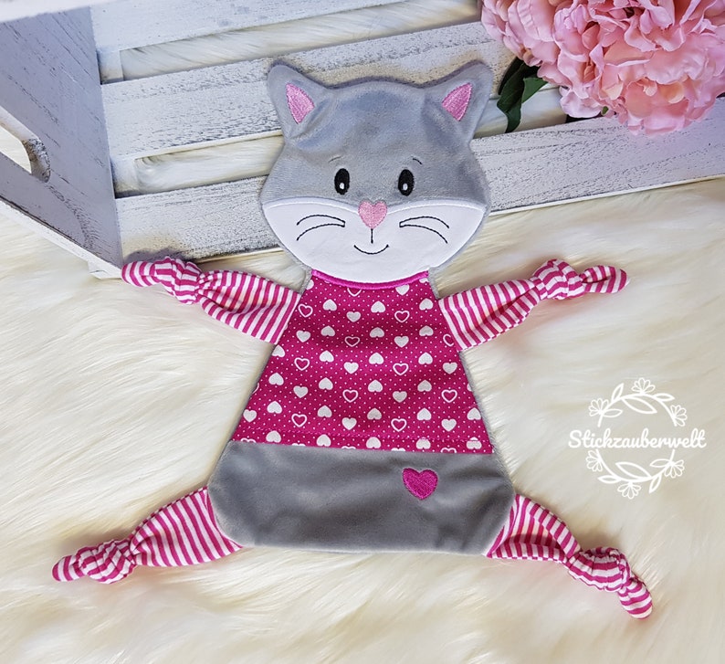 May include: A gray and pink cat-shaped plush toy with a pink heart pattern and white hearts. The toy has knotted arms and legs.