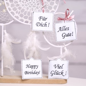 Peut inclure: Des attrape-rêves blancs avec des plumes et quatre cartes blanches avec du texte noir. Les cartes disent "Happy Birthday!" "Viel Glück!" "Alles Gute!" et "Für Dich!"