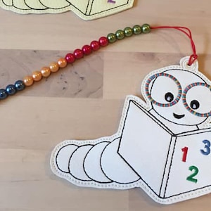 Könnte beinhalten: Ein skurriles Lesezeichen mit einem Cartoon-Wurm, der ein Buch mit den Zahlen 1, 2 und 3 liest. Das Lesezeichen hat eine Perlenkette mit blauen, orangefarbenen, roten und grünen Perlen.