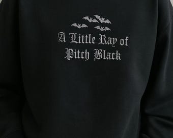 May include: Black sweatshirt with the phrase "A Little Ray of Pitch Black" in a gothic font, along with bat illustrations. The long-sleeved top is a solid black color.