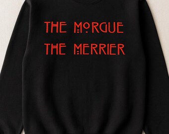 May include: Black crewneck sweatshirt with the red text "THE MORGUE THE MERRIER" in a bold, stylized font. The sweatshirt is a solid black color, and the text is centered on the chest.