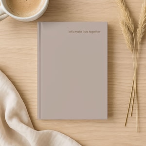 Puede incluir: Un cuaderno beige con la frase "let's make lists together". El cuaderno está sobre una superficie de madera clara, acompañado de una taza de café y hierba decorativa seca. El cuaderno es de un color suave y apagado.