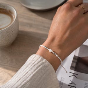 Könnte beinhalten: Weißes Kordelarmband mit silberfarbenen Perlen und einem kleinen herzförmigen Anhänger. Das Armband wird am Handgelenk getragen, im Hintergrund sind eine Kaffeetasse und eine Zeitschrift zu sehen.