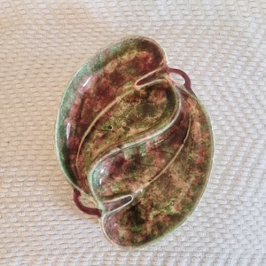 May include: A green and red ceramic dish shaped like a leaf with two handles. The dish has a mottled glaze.