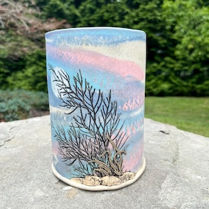 May include: A cylindrical ceramic vase with a pastel sky design in shades of blue, pink, and white. A black tree branch design is featured on the front, with a full moon. The vase is sitting on a gray stone surface.