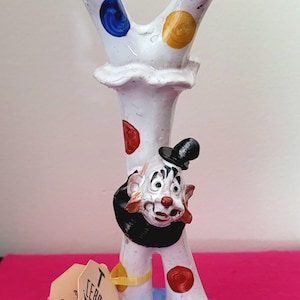 May include: White ceramic clown figurine with a black hat and red, yellow, and blue polka dots. The clown is standing on its hands with its legs in the air. The figurine is on a blue base with the text "TP Ceramiche" on a tag.