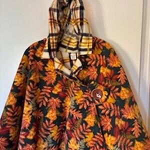 May include: A hooded cape with a fall leaf pattern in shades of orange, yellow, and red on a dark green background. The hood and collar feature a plaid design in similar colors. A large, round, brown button secures the cape.