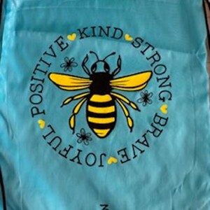 May include: A light blue drawstring bag featuring a yellow and black bee illustration within a circle. The circle includes the words "POSITIVE KIND STRONG BRAVE JOYFUL" and small heart and flower accents. The text "3 Sisters" is at the bottom.