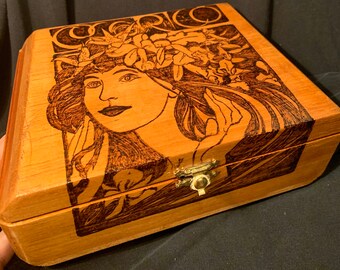 May include: A wooden box with a hinged lid, featuring a detailed pyrography design. The artwork depicts a woman with flowing hair and floral accents. The box has a brass clasp and a natural wood finish.