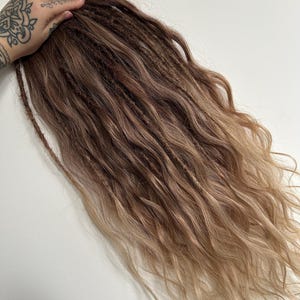 May include: A bundle of brown and blonde dreadlocks. The dreadlocks are a mix of dark brown and light blonde, with a wavy texture. The hair is styled to appear natural and flowing.