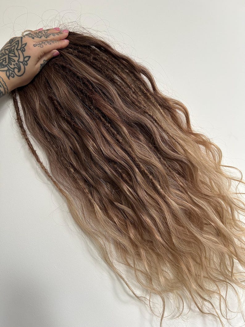 May include: A bundle of brown and blonde dreadlocks. The dreadlocks are a mix of dark brown and light blonde, with a wavy texture. The hair is styled to appear natural and flowing.