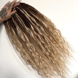 May include: A bundle of brown and blonde dreadlocks with wavy ends. The dreadlocks transition from a dark brown at the roots to a lighter blonde at the ends. The hair is styled with a natural, textured appearance.