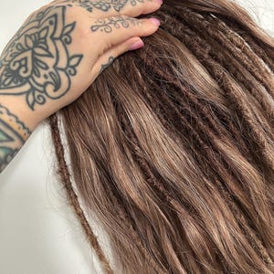 May include: Brown and light brown dreadlocks, with a natural, textured appearance. The dreadlocks are shown against a white background. The image shows the dreadlocks in a close-up view.