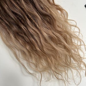 May include: Close-up of long, wavy hair extensions in shades of brown and blonde. The hair is styled in dreadlocks, with a mix of textures and colors. The ends of the hair are lighter, creating a natural ombre effect.