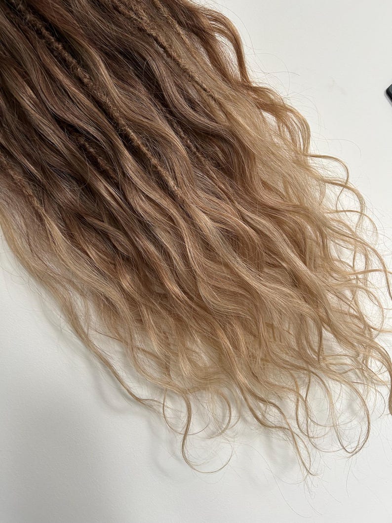 May include: Close-up of long, wavy hair extensions in shades of brown and blonde. The hair is styled in dreadlocks, with a mix of textures and colors. The ends of the hair are lighter, creating a natural ombre effect.