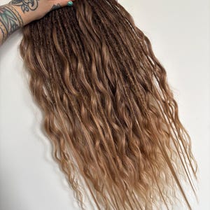 May include: A bundle of brown and blonde dreadlocks with a wavy texture. The dreadlocks are tied together with a brown ribbon. The hair transitions from a darker brown at the top to a lighter blonde at the ends.