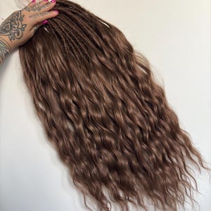 Pode incluir: Extensões de cabelo castanho ondulado. As extensões são de uma cor castanha quente com uma mistura de secções onduladas e trançadas. O cabelo é estilizado em longas ondas fluidas.