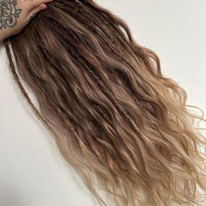 Könnte beinhalten: Ein Bündel brauner und blond gefärbter Dreadlocks mit welliger Textur. Die Haarverlängerungen haben einen Farbverlauf, der von einem dunkleren Braun an den Wurzeln zu einem helleren Blond an den Enden übergeht. Die Dreadlocks werden von einer Hand mit Tätowierung gehalten.
