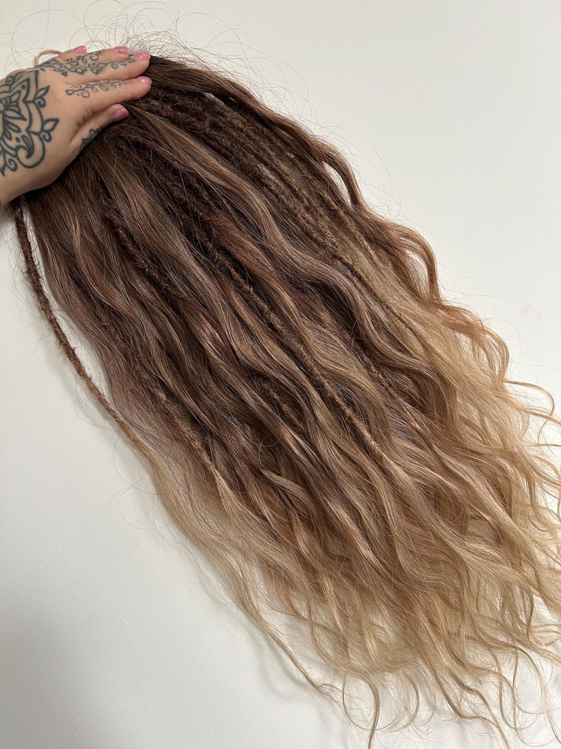 May include: A bundle of brown and blonde dreadlocks with a wavy texture. The hair extensions have a gradient effect, transitioning from a darker brown at the roots to a lighter blonde at the ends. The dreadlocks are held by a hand with a tattoo.