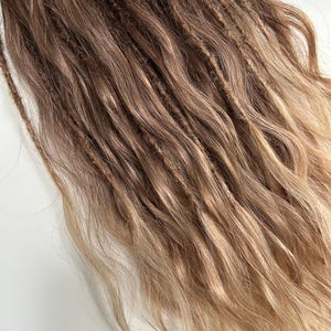 May include: A close-up of brown and blonde dreadlocks. The dreadlocks are a mix of light and dark brown, with blonde highlights at the ends. The hair is wavy and textured, with a natural look.