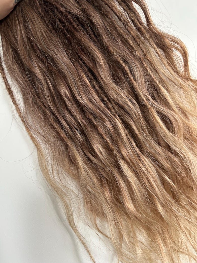 May include: A close-up of brown and blonde dreadlocks. The dreadlocks are a mix of light and dark brown, with blonde highlights at the ends. The hair is wavy and textured, with a natural look.