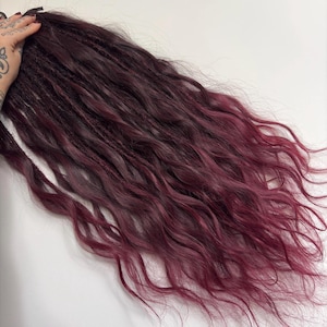 Puede incluir: Dreadlocks de color burdeos oscuro con puntas onduladas. Las extensiones de cabello hacen la transición de un color profundo y rico en las raíces a un tono vibrante y más claro en las puntas. Los dreadlocks tienen un aspecto natural y fluido.