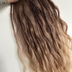 May include: A bundle of brown and blonde dreadlocks with a wavy texture. The dreadlocks are a mix of dark brown at the roots, transitioning to lighter brown and blonde at the ends. The hair is styled to appear natural.