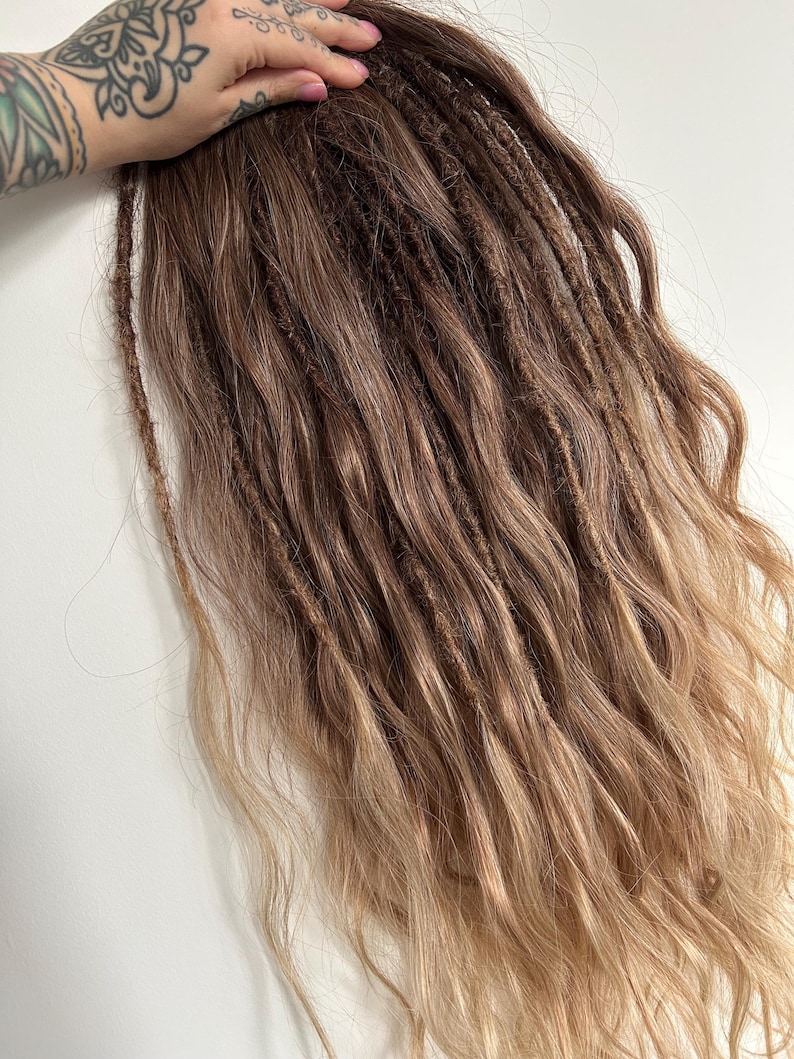 May include: A bundle of brown and blonde dreadlocks with a wavy texture. The dreadlocks are a mix of dark brown at the roots, transitioning to lighter brown and blonde at the ends. The hair is styled to appear natural.