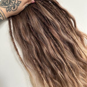 May include: A bundle of brown and blonde dreadlocks. The dreadlocks are a mix of dark brown and lighter blonde shades, creating a natural, blended look. The hair is styled in individual dreads, with a textured appearance. The ends of the hair have a lighter, sun-kissed effect.