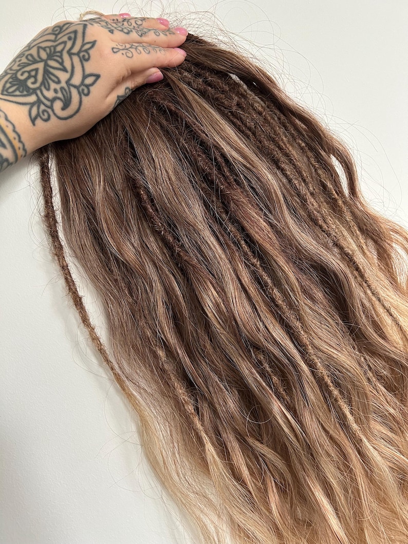 May include: A bundle of brown and blonde dreadlocks. The dreadlocks are a mix of dark brown and lighter blonde shades, creating a natural, blended look. The hair is styled in individual dreads, with a textured appearance. The ends of the hair have a lighter, sun-kissed effect.
