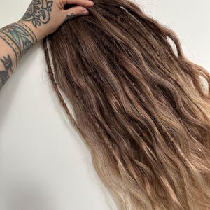 May include: A bundle of brown and blonde dreadlocks. The dreadlocks are a mix of dark brown and lighter blonde shades, with a natural, textured appearance. The hair extensions are styled in a wavy pattern.