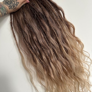 May include: Brown and blonde dreadlocks with wavy ends. The hair extensions transition from a dark brown at the roots to a lighter blonde at the tips. The dreadlocks are styled with a textured appearance.
