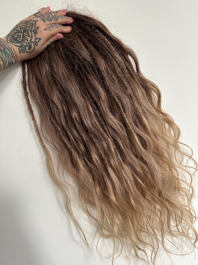May include: Brown and blonde dreadlocks with wavy ends. The hair extensions transition from a dark brown at the roots to a lighter blonde at the tips. The dreadlocks are styled with a textured appearance.