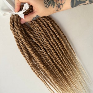 Puede incluir: Un paquete de extensiones de cabello largas y ombre. Las extensiones son de color marrón claro en la parte superior y se desvanecen a un rubio claro en los extremos. Las extensiones están trenzadas en una trenza con forma de cuerda.