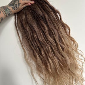 May include: A bundle of brown and blonde dreadlocks with a wavy texture. The dreadlocks are held by a hand with tattoos. The hair transitions from a darker brown at the roots to a lighter blonde at the ends.