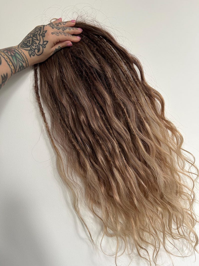 May include: A bundle of brown and blonde dreadlocks with a wavy texture. The dreadlocks are held by a hand with tattoos. The hair transitions from a darker brown at the roots to a lighter blonde at the ends.
