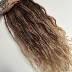 May include: A bundle of brown and blonde dreadlocks with wavy ends. The dreadlocks are a mix of dark brown at the roots, transitioning to lighter blonde at the tips. The hair is styled with a natural, wavy texture.