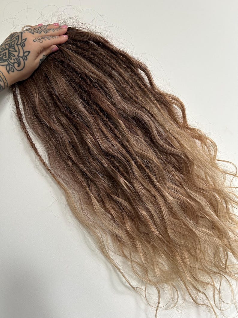 May include: A bundle of brown and blonde dreadlocks with wavy ends. The dreadlocks are a mix of dark brown at the roots, transitioning to lighter blonde at the tips. The hair is styled with a natural, wavy texture.