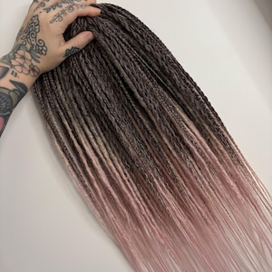 May include: A bundle of braided hair extensions with a color gradient. The top portion is a dark brown, transitioning to a lighter brown, and finally fading into a soft pink at the ends. The hair is held by a hand with tattoos.