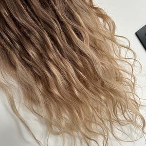 May include: Close-up of a set of dreadlocks with a blend of brown and blonde tones. The dreads have a textured appearance, with some sections appearing more tightly wound than others. The ends of the hair transition to a lighter, wavy blonde.
