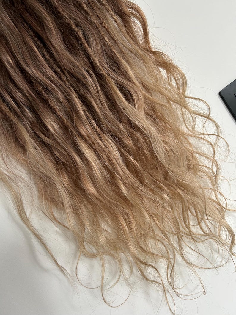 May include: Close-up of a set of dreadlocks with a blend of brown and blonde tones. The dreads have a textured appearance, with some sections appearing more tightly wound than others. The ends of the hair transition to a lighter, wavy blonde.