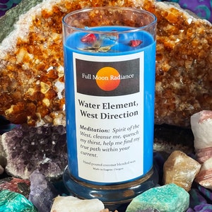 May include: A blue glass candle with a white label that reads "Full Moon Radiance, Water Element, West Direction". The label also includes a meditation message and details about the candle being hand-poured coconut blended wax made in Eugene, Oregon.