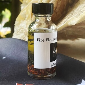 May include: A small glass bottle with a black cap filled with a clear liquid and brown herbs. The label reads "Fire Element" and "Full Moon Magic".