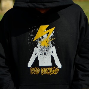 Può includere: Felpa con cappuccio nera con una stampa grafica bianca e gialla di una persona con un fulmine nella testa. Il testo "BAD BRAINS" è stampato sotto la grafica.